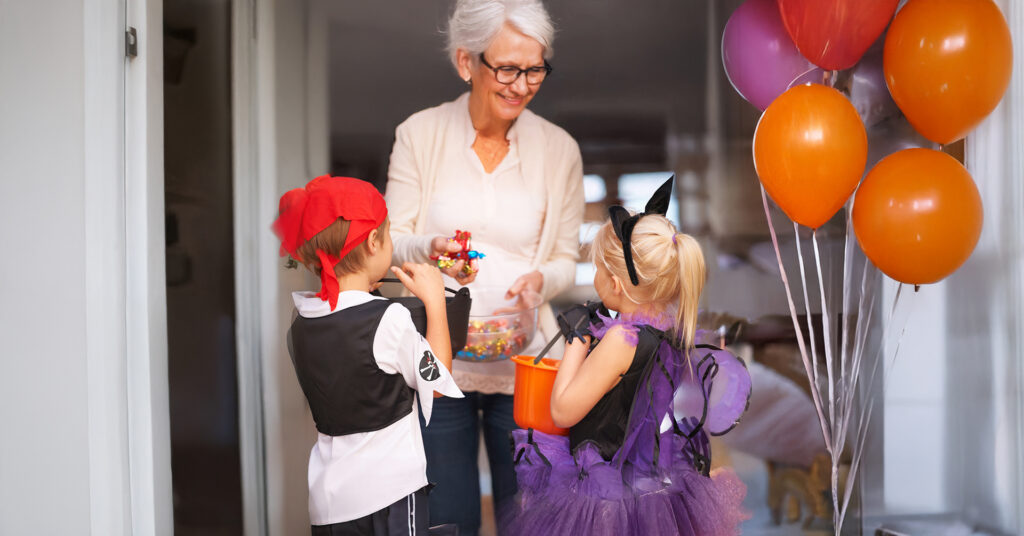 senior woman handing out candy to 2 children trick-or-treating dressed up in costumes for Halloween