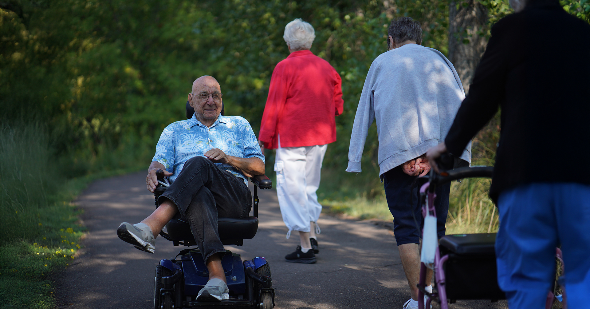 senior man enjoying a sunny day on a paved walking path in his wheelchair passing a group of older ladies using a walker