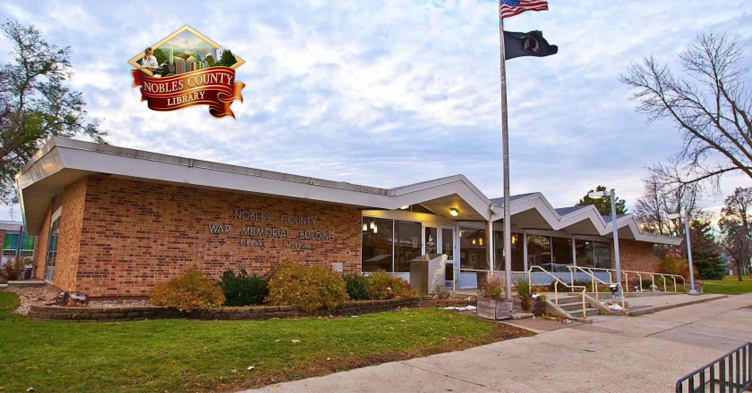 Exterior of a brick library with zigzag roofline, surrounded by greenery. Flags on poles, with an open, welcoming entrance under a cloudy sky. Logo in upper-left corner reads "Nobles County Library."