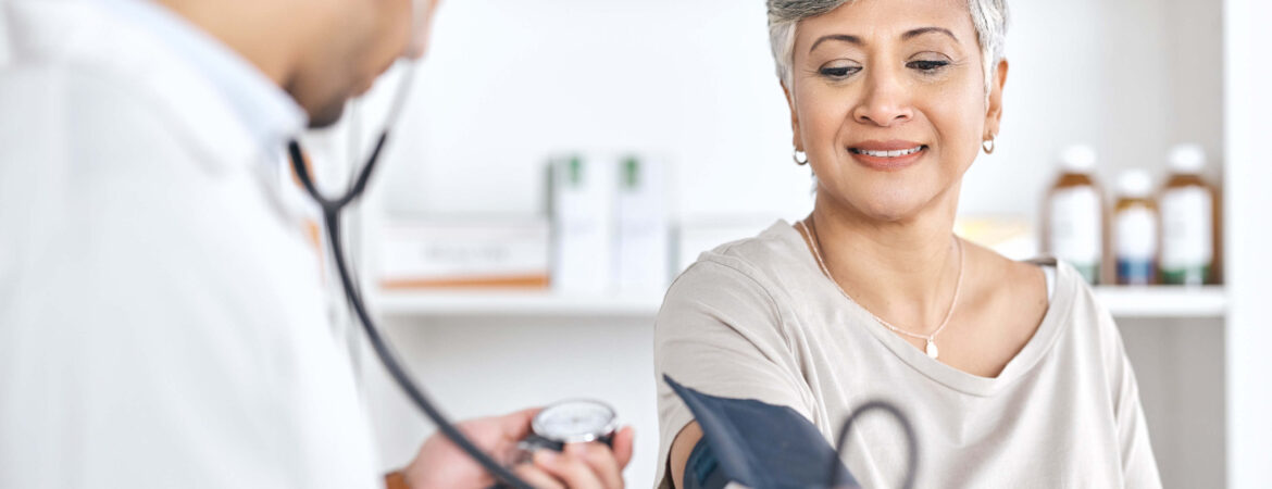 A healthcare professional measures a smiling woman's blood pressure in a clinic. The calm, friendly atmosphere conveys trust and care.