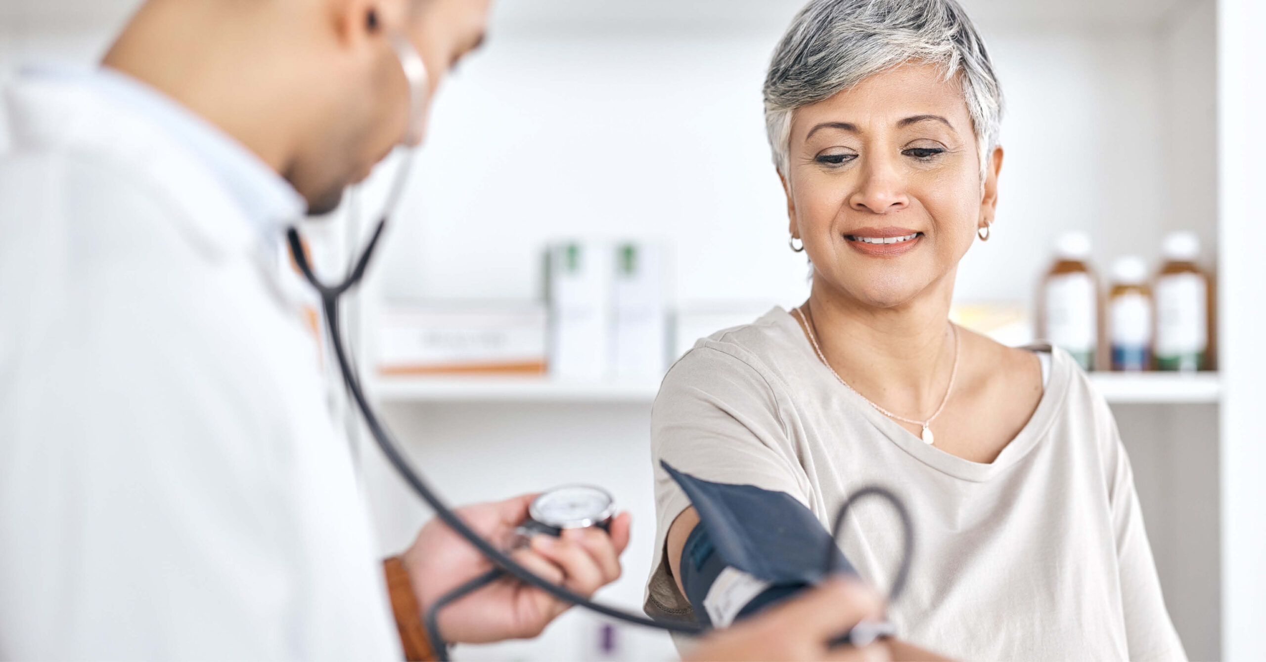A healthcare professional measures a smiling woman's blood pressure in a clinic. The calm, friendly atmosphere conveys trust and care.
