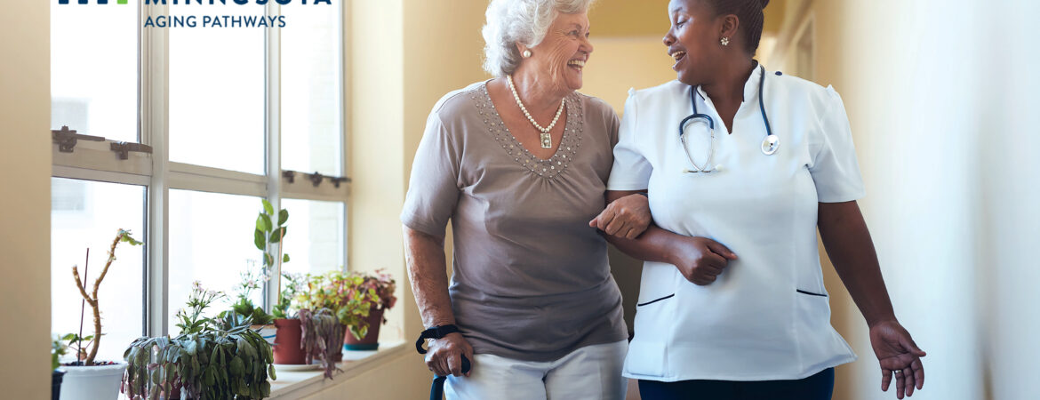 A nurse and an older woman walk arm-in-arm, smiling in a sunlit room with plants.