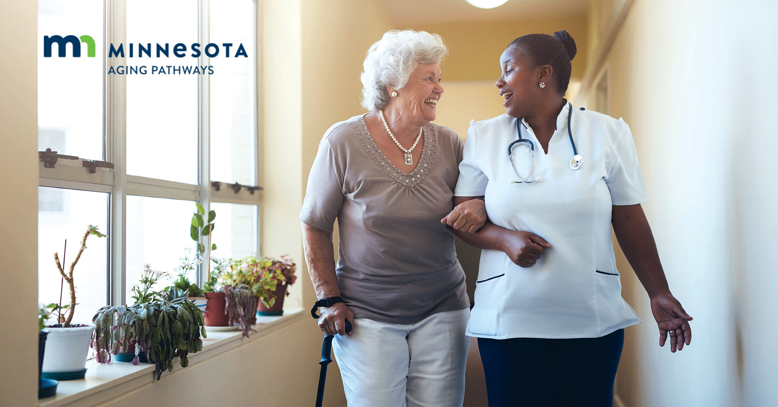A nurse and an older woman walk arm-in-arm, smiling in a sunlit room with plants.