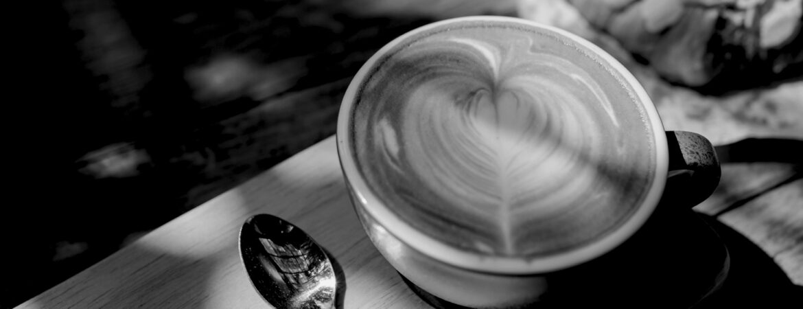 A black and white photo of a cup of latte art and a silver spoon.