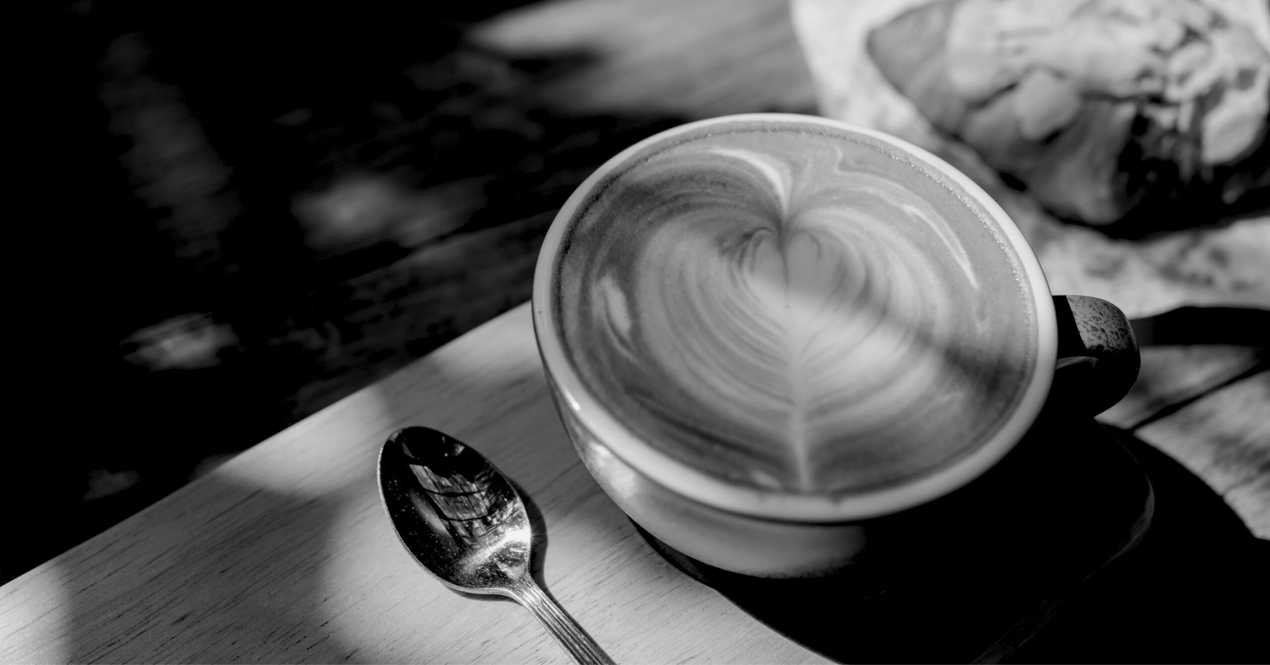 A black and white photo of a cup of latte art and a silver spoon.